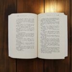 An open paperback book laid flat on a wooden table, pages spread with visible text in two columns on each page edge-to-edge.