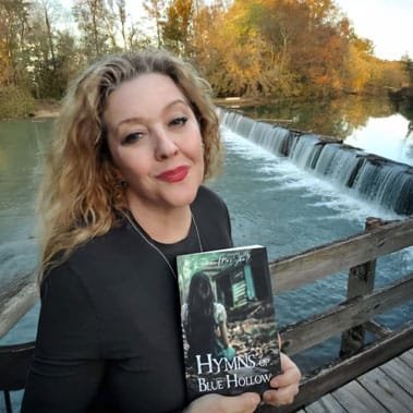 Author Kemma MarShall holding a copy of Hymns of Blue Hollow beside a river waterfall in autumn
